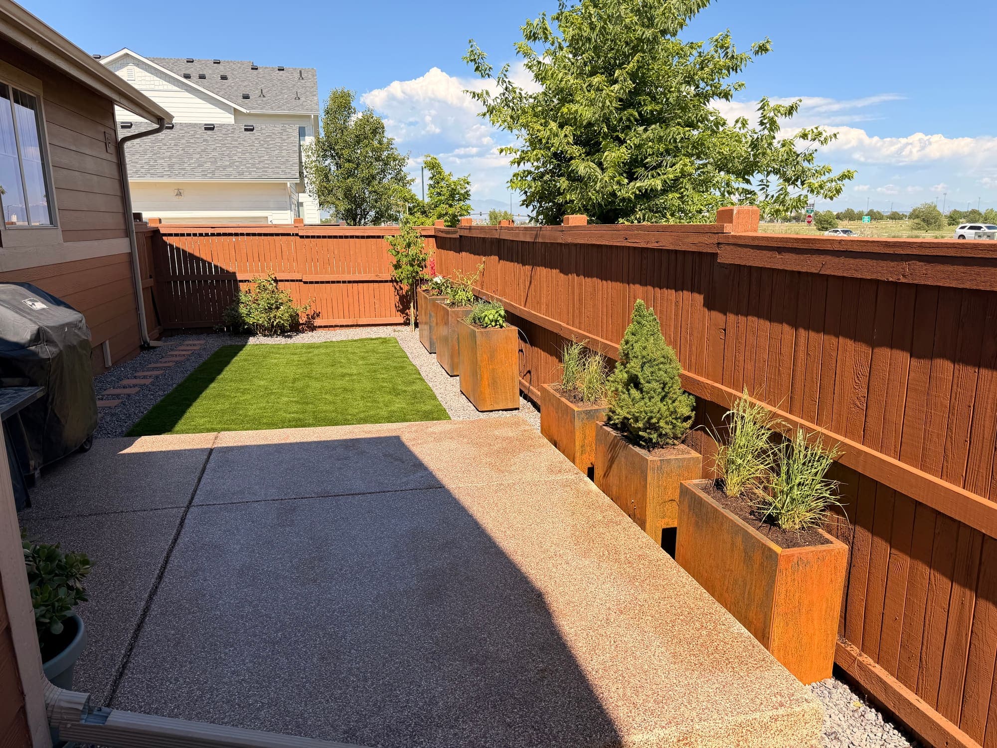 The same backyard after: turf lawn, exposed-aggregate patio, and corten steel planters of grasses and evergreens along the fence